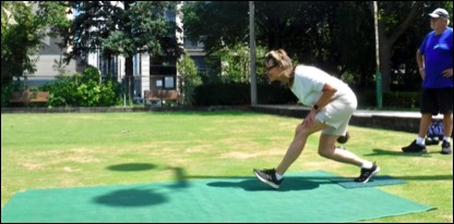 Female lawn bowler delivering a bowl with a male lawn bowler watching in the background.